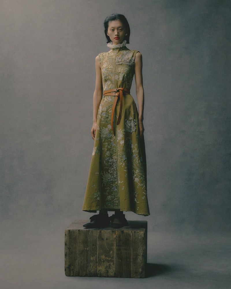 Asian female model in a sleeveless green floral dress with a brown belt stands on a wooden crate against a grey studio backdrop.