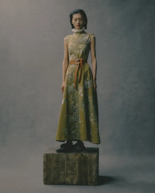 Asian female model in a sleeveless green floral dress with a brown belt stands on a wooden crate against a grey studio backdrop.
