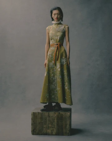 Asian female model in a sleeveless green floral dress with a brown belt stands on a wooden crate against a grey studio backdrop.