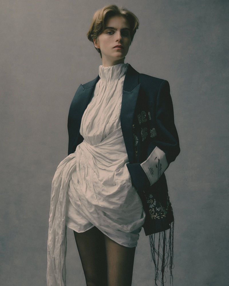 Female model in a draped white dress and navy blazer poses indoors against a grey background in a fashion editorial shot.