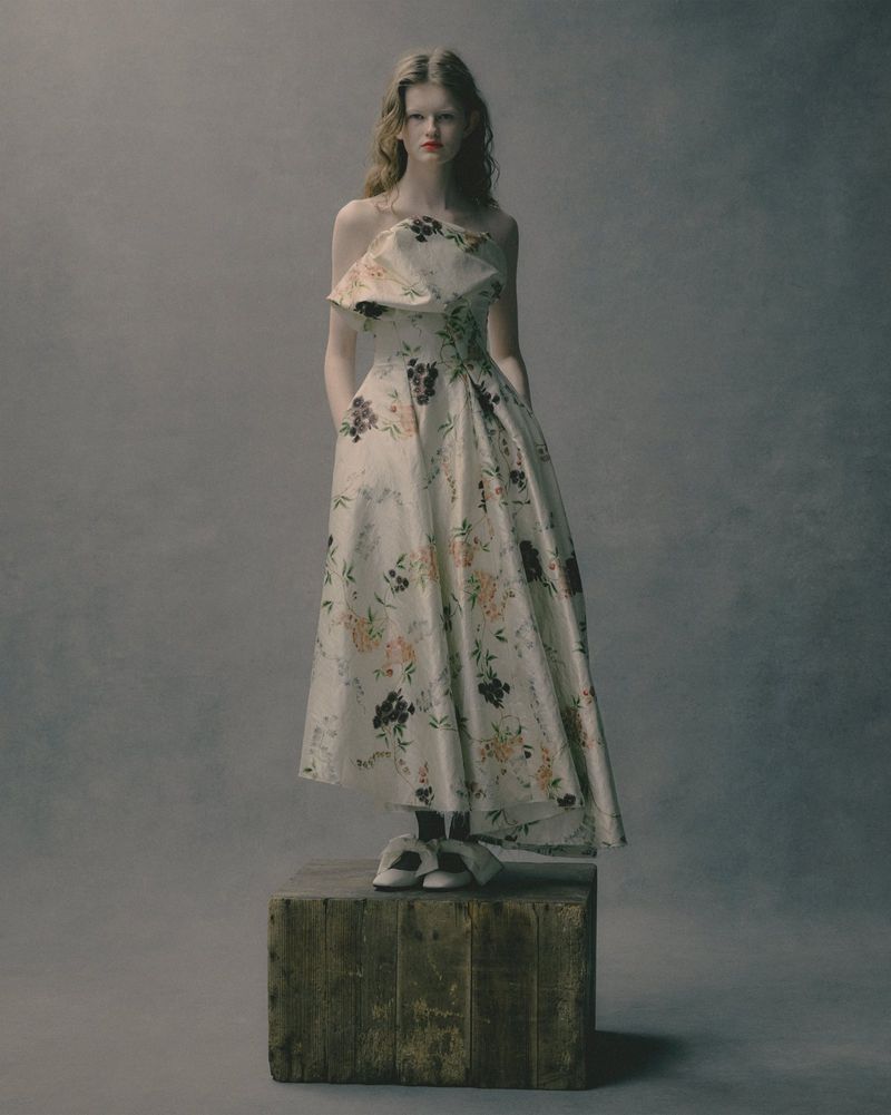 Woman in floral off-the-shoulder dress standing on a wooden crate in a studio. The background is a soft grey.