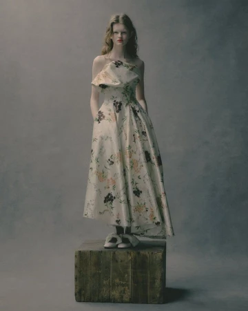 Woman in floral off-the-shoulder dress standing on a wooden crate in a studio. The background is a soft grey.