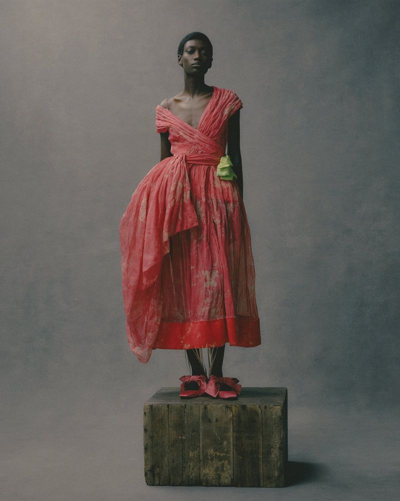 Model wearing a draped pink dress standing on a weathered wooden crate against a dark studio background