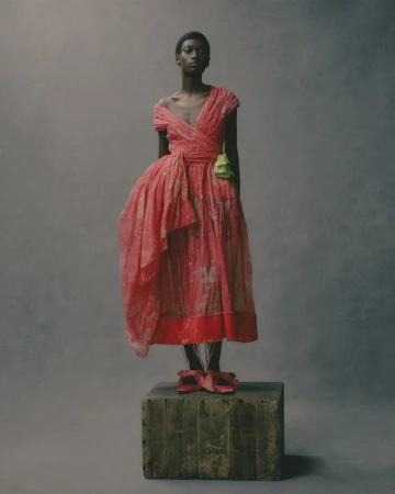 Model wearing a draped pink dress standing on a weathered wooden crate against a dark studio background