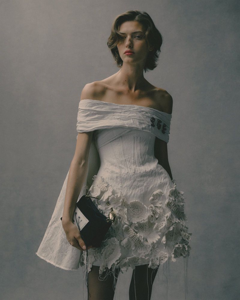 Model in off-the-shoulder white dress holding black handbag against textured grey backdrop for Erdem S/S 26
