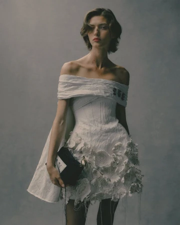 Model in off-the-shoulder white dress holding black handbag against textured grey backdrop for Erdem S/S 26