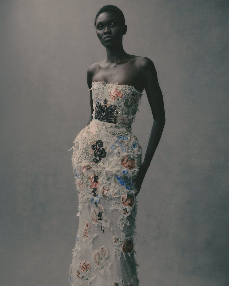 Woman in a floor-length strapless gown with floral embellishments poses in a studio with a neutral background.
