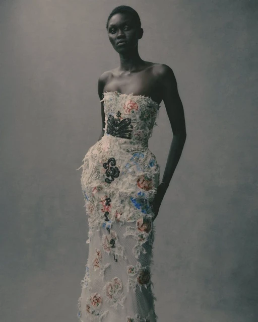 Woman in a floor-length strapless gown with floral embellishments poses in a studio with a neutral background.