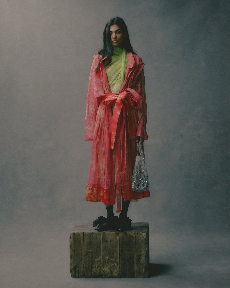 A full-length studio portrait of a woman in a pink robe standing on a wooden crate holding a silver beaded purse.