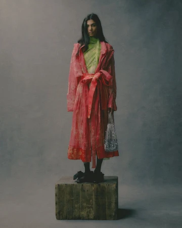 A full-length studio portrait of a woman in a pink robe standing on a wooden crate holding a silver beaded purse.