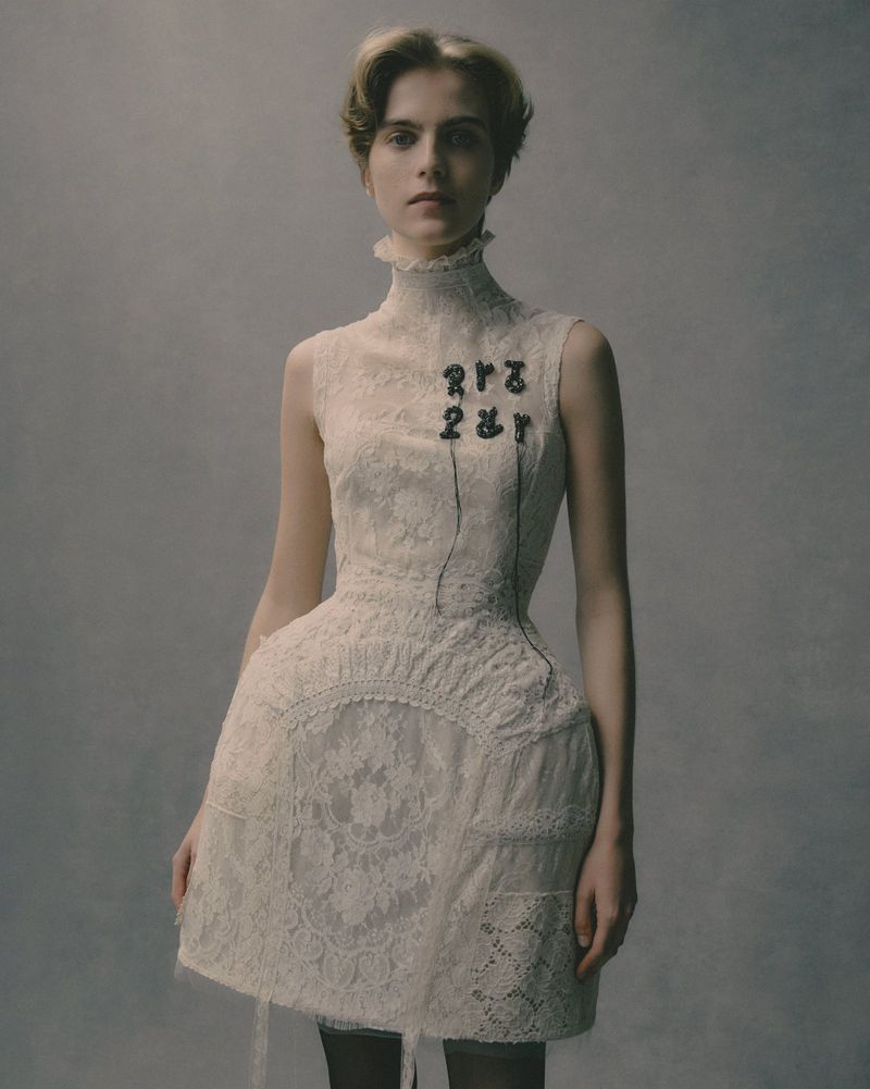 A fair-skinned young woman poses in a high-necked, sleeveless white lace dress against a diffused grey background in a fashion shot.