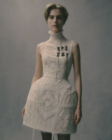 A fair-skinned young woman poses in a high-necked, sleeveless white lace dress against a diffused grey background in a fashion shot.