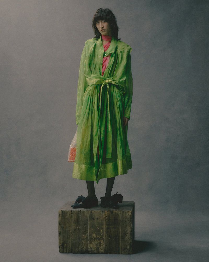 Full shot of a young Asian woman in a green sheer dress standing on a wood crate in a studio with a neutral grey backdrop.