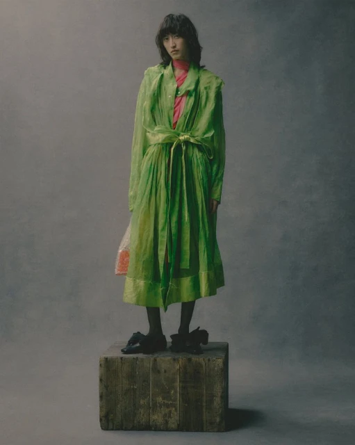 Full shot of a young Asian woman in a green sheer dress standing on a wood crate in a studio with a neutral grey backdrop.