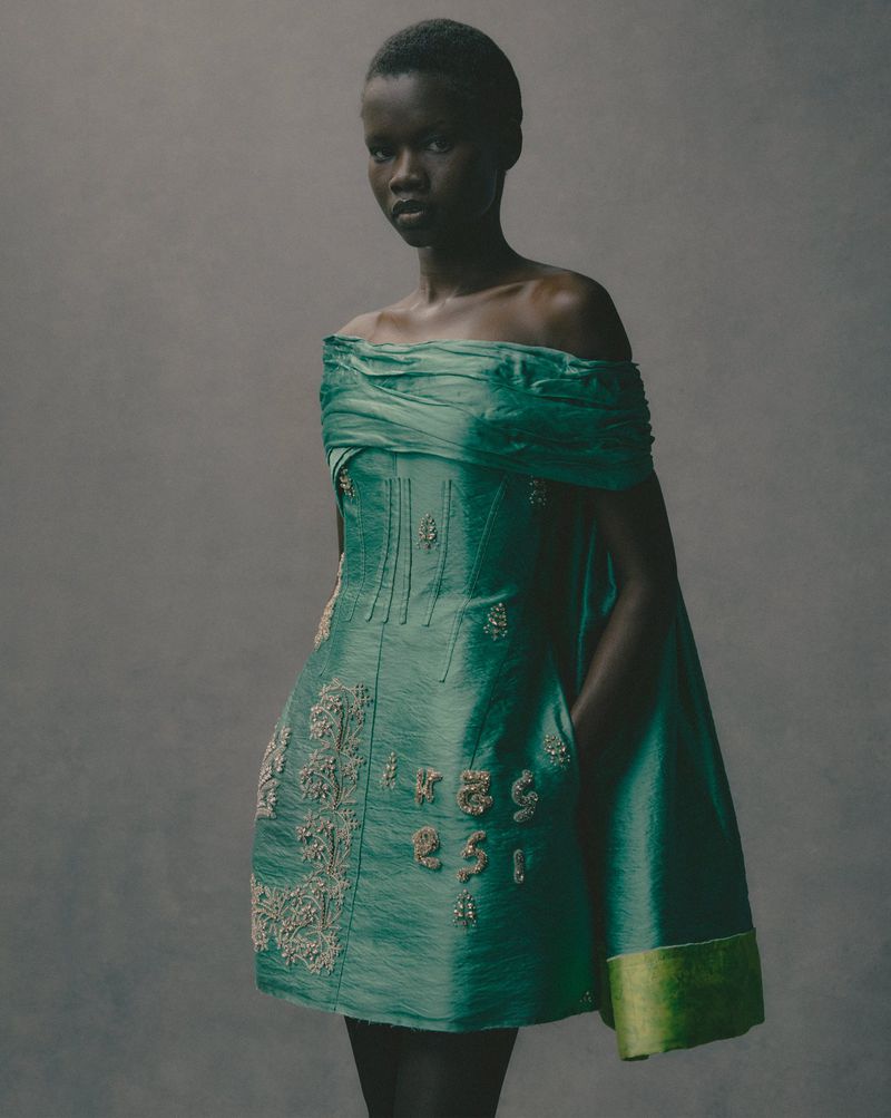 Portrait of a Black woman wearing a green off-shoulder dress with embellishments, posing against a neutral-toned backdrop in studio lighting.