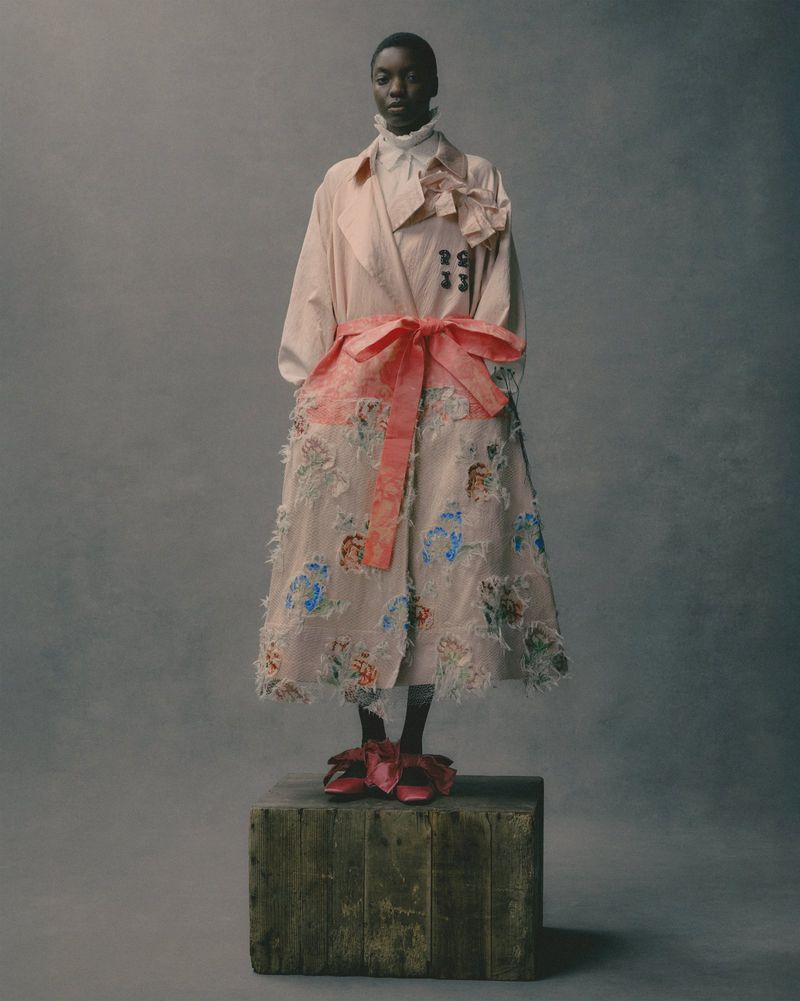 An African-American woman poses on a wooden box in a pink floral trench coat and matching flats against a muted grey studio backdrop.
