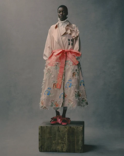 An African-American woman poses on a wooden box in a pink floral trench coat and matching flats against a muted grey studio backdrop.