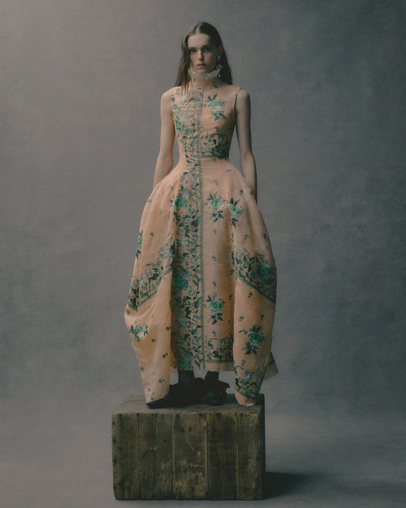 Fashionable adult woman in a peach gown with floral detail standing on a wooden box in a studio with a muted grey backdrop.