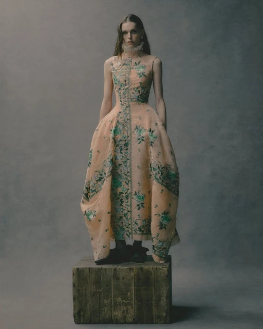 Fashionable adult woman in a peach gown with floral detail standing on a wooden box in a studio with a muted grey backdrop.
