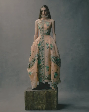 Fashionable adult woman in a peach gown with floral detail standing on a wooden box in a studio with a muted grey backdrop.