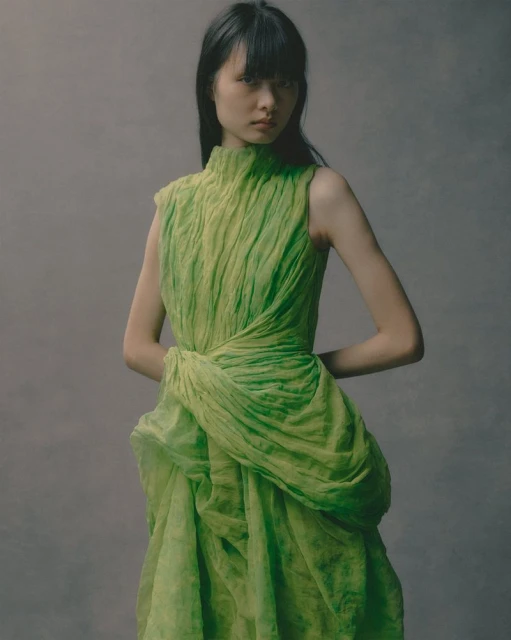 A striking portrait captures a slender Asian woman in a green textured gown, striking an elegant pose against a soft grey backdrop.