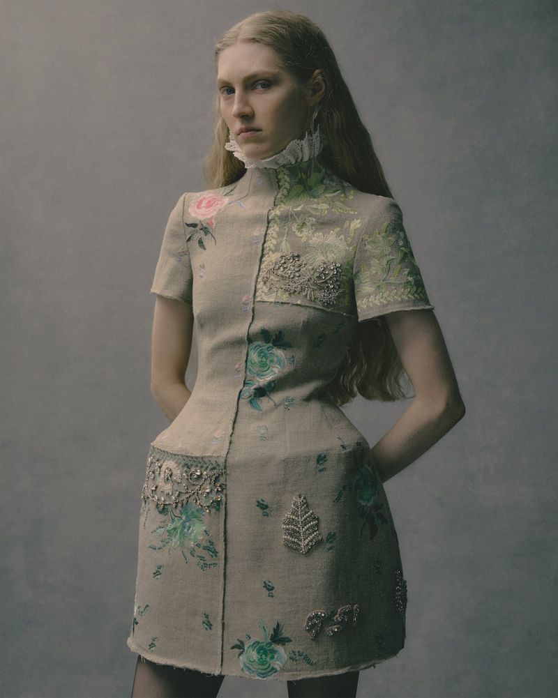 Woman in a floral-embellished dress with a high ruffled collar, posing against a neutral grey backdrop in a fashion studio shoot.