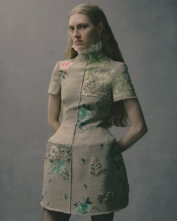 Woman in a floral-embellished dress with a high ruffled collar, posing against a neutral grey backdrop in a fashion studio shoot.