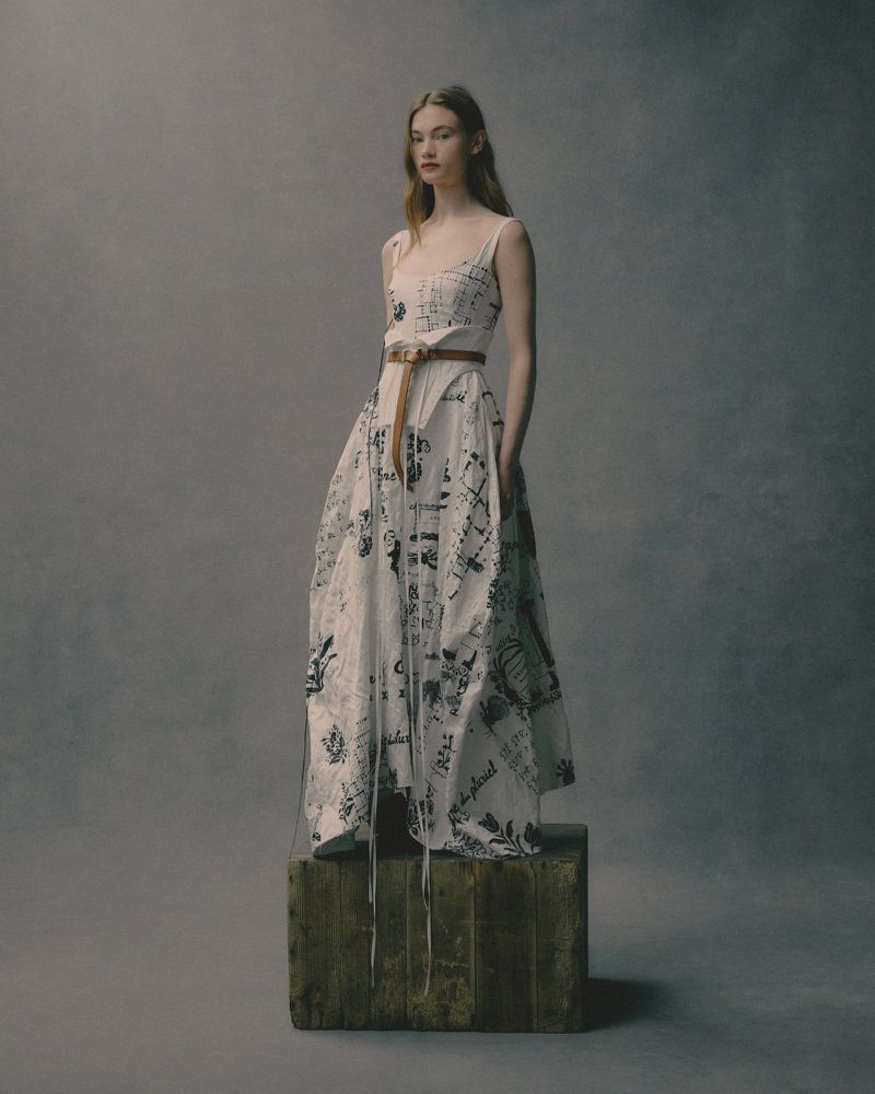 Full shot of a woman in a patterned dress posing on a wooden cube against a gray background in a fashion studio setting.