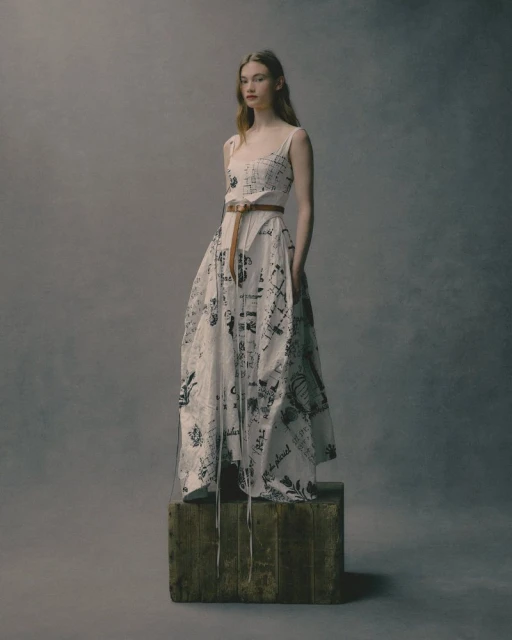 Full shot of a woman in a patterned dress posing on a wooden cube against a gray background in a fashion studio setting.