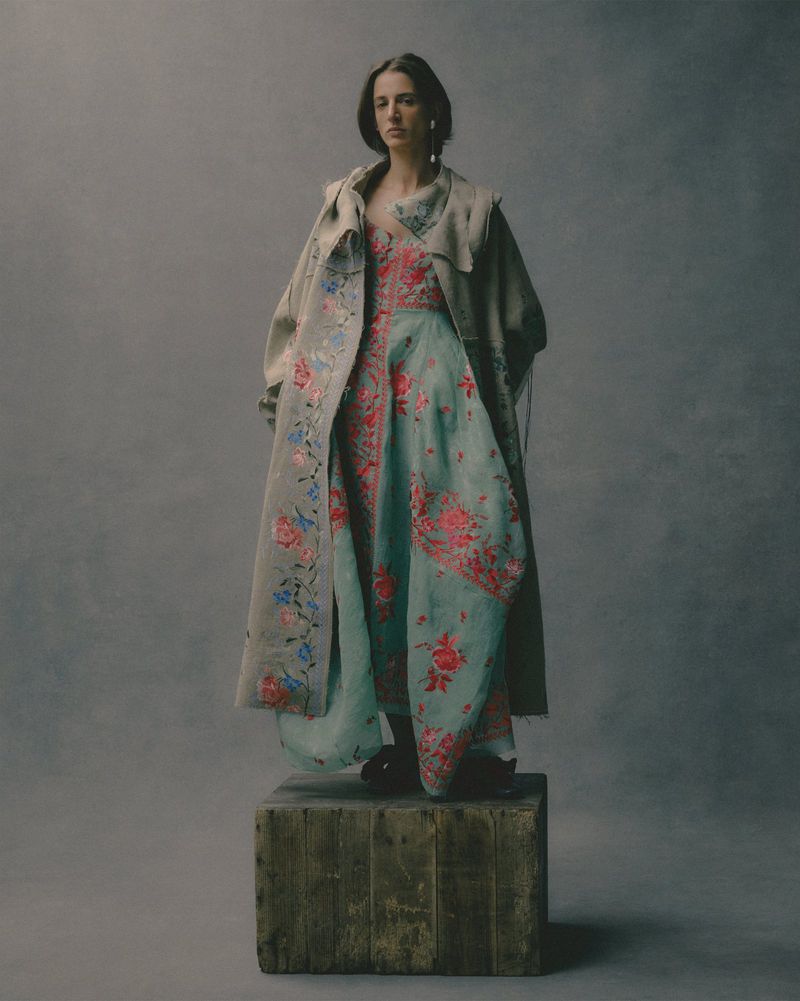 Model standing on a weathered wooden crate in a floral dress and draped coat against a textured grey studio background.