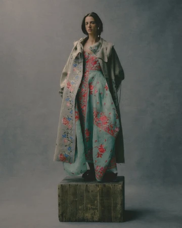 Model standing on a weathered wooden crate in a floral dress and draped coat against a textured grey studio background.