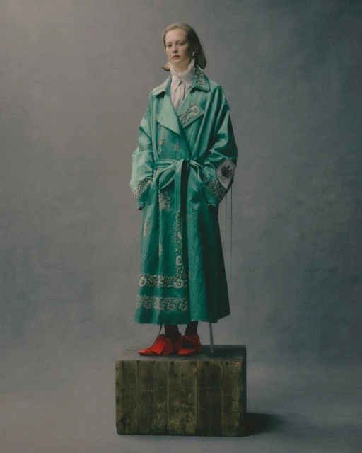 Full-length portrait of a fair-skinned woman in a teal embroidered trench coat, posing on a wooden box against a neutral gray background.