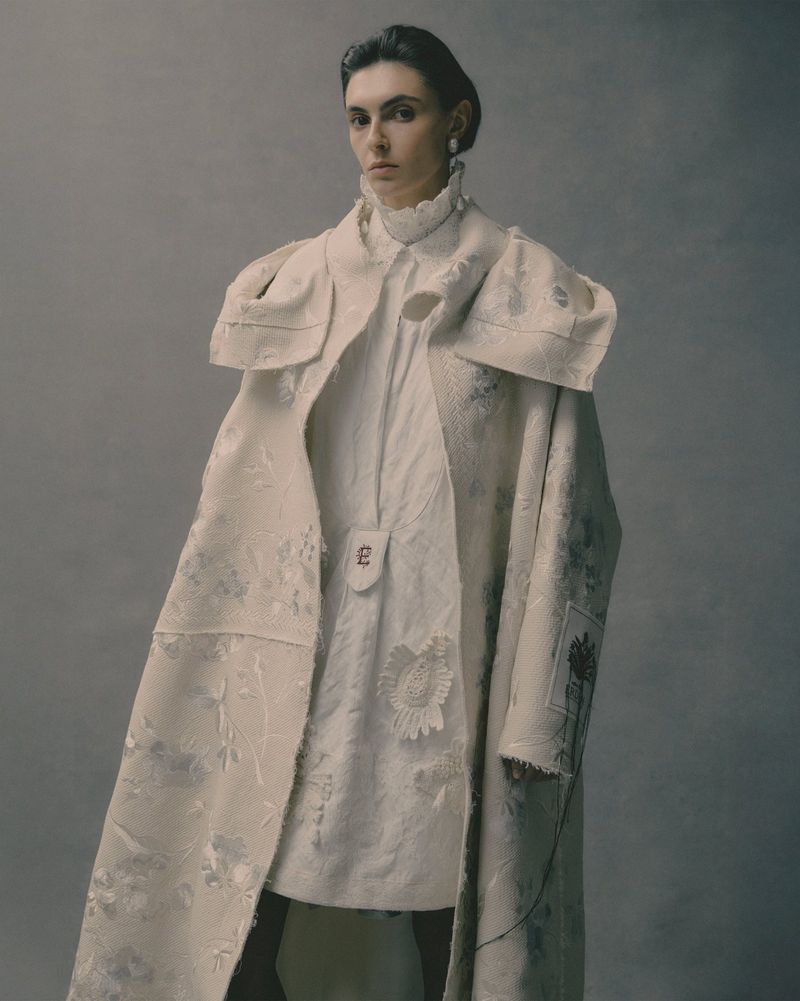 Model wearing a textured cream coat and ruffled collar dress against a neutral grey studio backdrop.