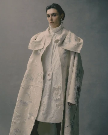 Model wearing a textured cream coat and ruffled collar dress against a neutral grey studio backdrop.