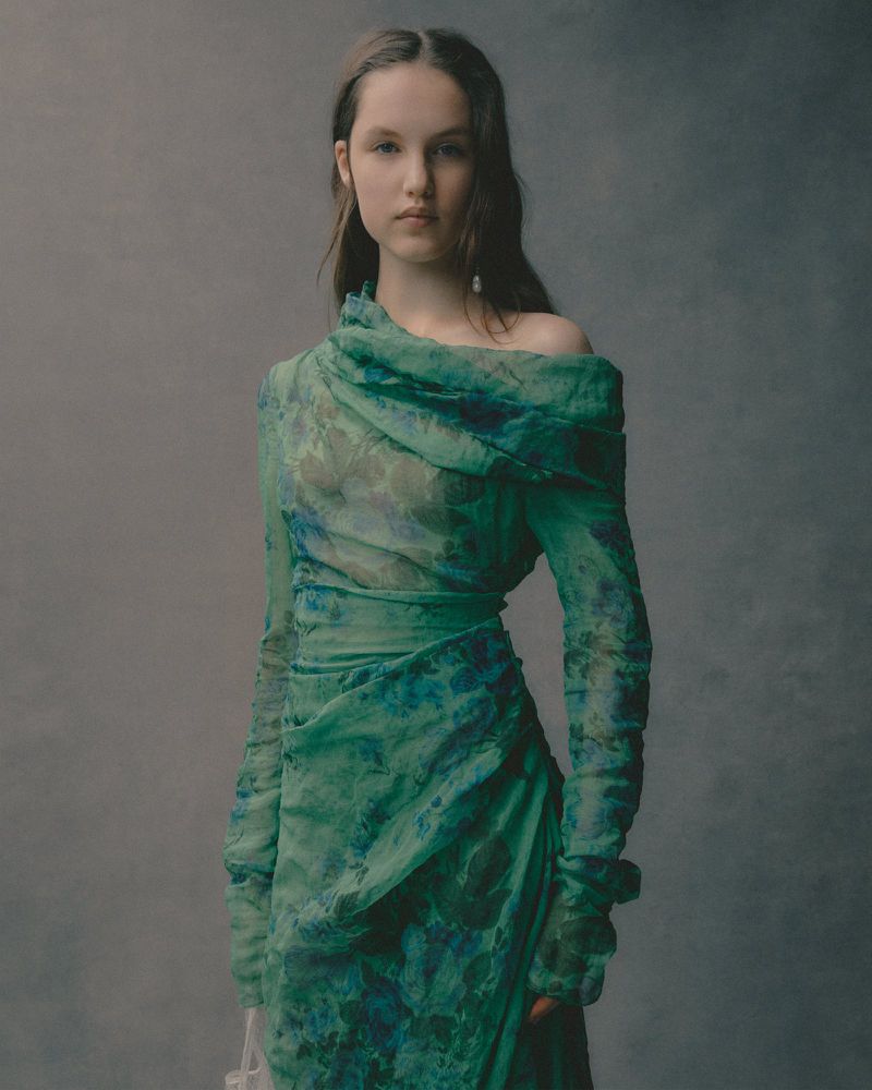 Elegant woman in a flowing green and blue floral gown, posing against a soft grey backdrop, fashion shoot, long sleeves and draping.