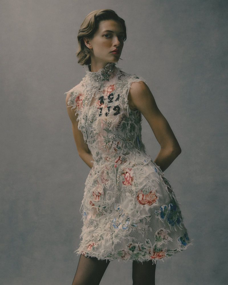 Woman in embellished floral dress and black tights poses in a studio with soft lighting against a solid grey backdrop for a fashion shot.