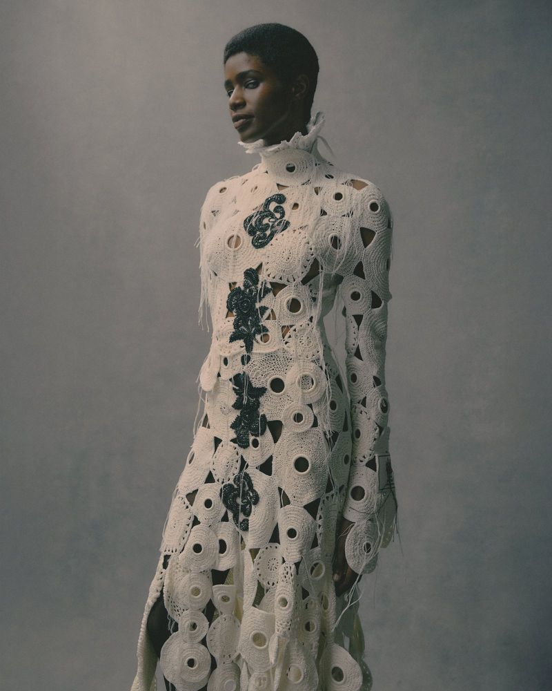 Fashion shot of a Black female model wearing a long, off-white, semi-sheer dress with decorative black floral appliques, studio setting.