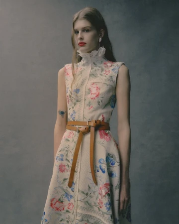 Model in a floral print high-neck dress with a thin brown leather belt against a textured grey studio background