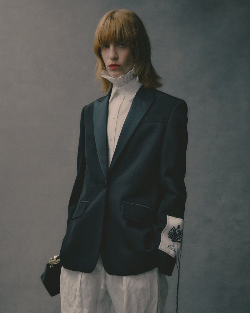 Model with reddish blonde hair wearing a dark blazer, ruffled white shirt, and white trousers against a grey backdrop