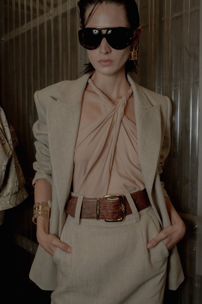An adult woman poses in a beige skirt suit with a brown belt, accessorized with gold jewelry and aviator sunglasses, against a dark background.