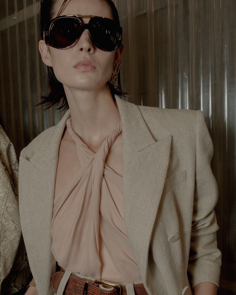 Fashion portrait of a woman wearing sunglasses and a tan blazer in a neutral, indoor setting. The lighting is soft and diffused.