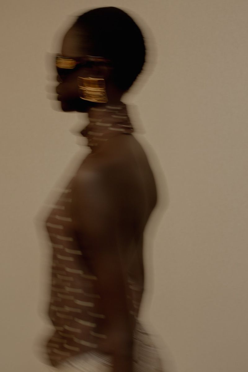 Side profile shot of a dark-skinned adult female with gold sunglasses and large gold square earrings against a white background.