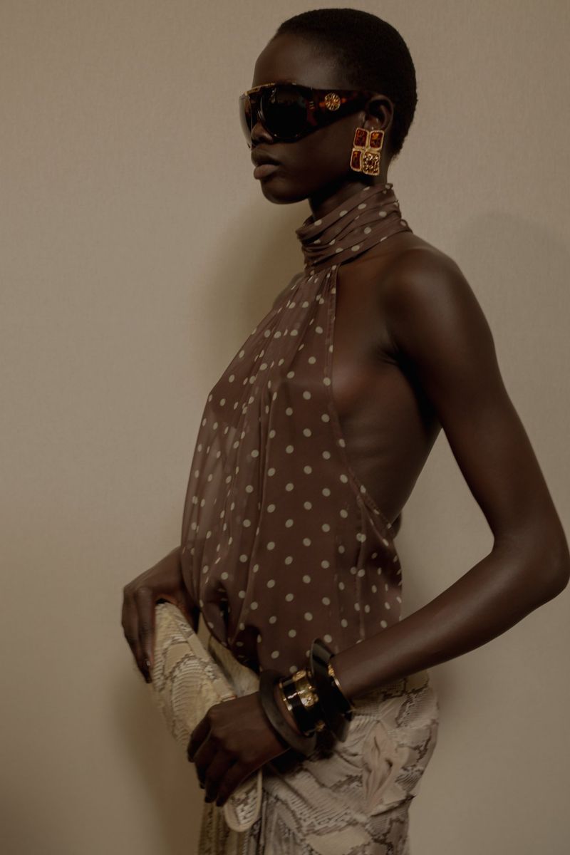 Elegant woman with dark skin in brown polka dot halter top, sunglasses, gold earrings, and snakeskin clutch poses in studio.