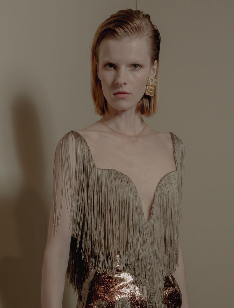 Close-up of a blonde woman with short hair wearing a dress with gold fringe and large gold earrings, posing indoors against a neutral background.