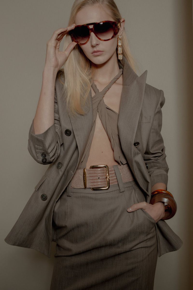 A fair-skinned woman in oversized sunglasses and a tailored grey-brown pantsuit, poses indoors for a fashion editorial photo.
