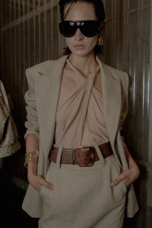 Model wearing dark sunglasses, a beige blazer, and a halter top with a wide brown belt against a corrugated wall.