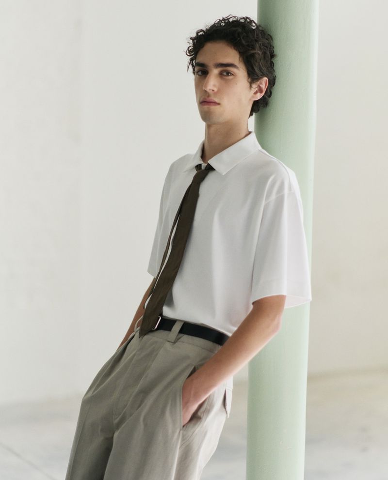 Studio portrait of a dark-haired man wearing a white polo shirt and tie, with khaki pants, leaning against a light-green column.