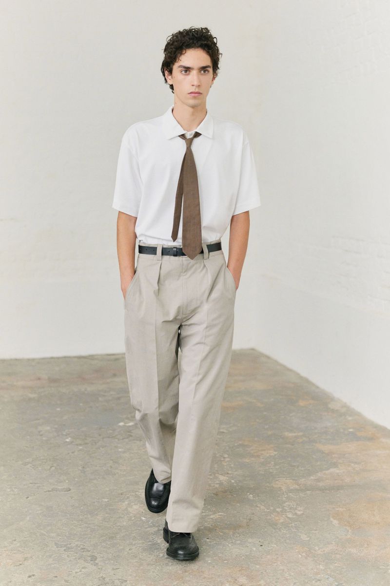 A young adult male walks forward wearing a white short-sleeved shirt, tan pants, brown tie, black belt, and black shoes in a studio setting.
