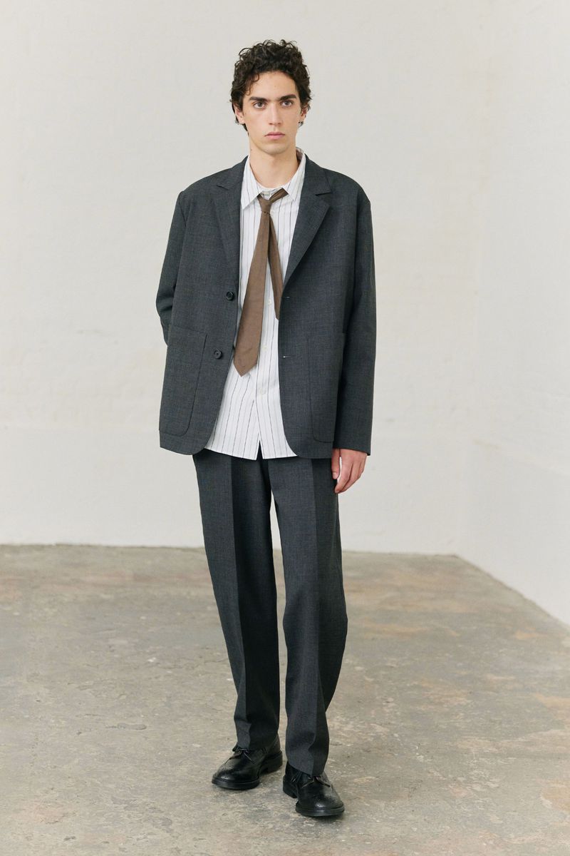 Male fashion model in a gray suit, white shirt, and brown tie poses in an indoor setting against a white wall and gray floor.