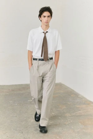Model wearing white short sleeve shirt, brown tie, and light grey trousers in a sparse indoor studio setting